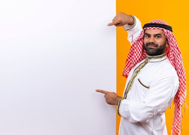 Arabische zakenman in traditionele slijtage die op leeg aanplakbord richt dat zich zelfverzekerd over oranje muur bevindt