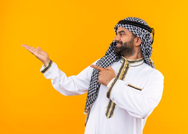 Arabische zakenman in traditionele slijtage die opzij kijkt wijzend met vinger en arm naar de zijkant staande over oranje muur