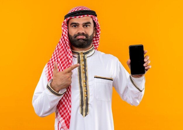 Arabische zakenman in traditionele slijtage die smartphone toont die met vinger ernaar richt kijkt die verward over oranje muur staat