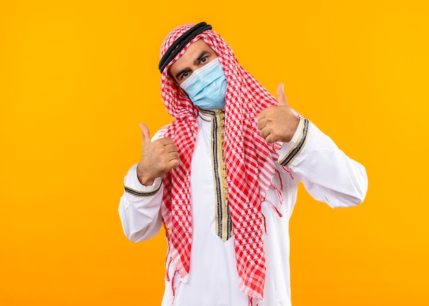 Arabische zakenman in traditionele slijtage en gezichts beschermend masker met zelfverzekerde uitdrukking die duimen toont die zich over oranje muur bevinden