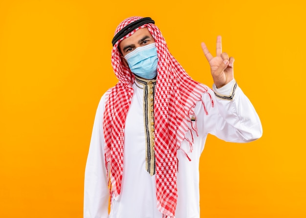 Arabische zakenman in traditionele slijtage en gezichts beschermend masker met zelfverzekerde uitdrukking die overwinningsteken toont dat zich over oranje muur bevindt
