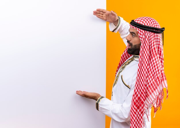 Arabische zakenman in traditionele slijtage wijzend op leeg bord met armen kijkt verbaasd staande over oranje muur