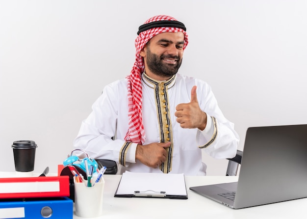 Arabische zakenman in traditionele slijtage zittend aan de tafel met laptop computer blij en positief glimlachen duimen opdagen werken in kantoor