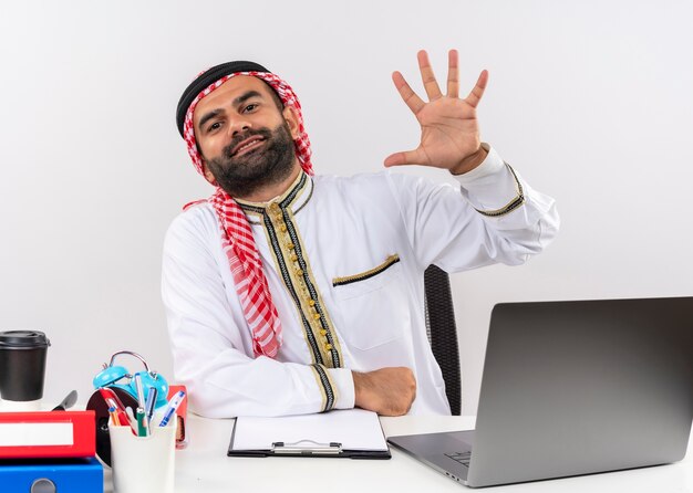 Arabische zakenman in traditionele slijtage zittend aan de tafel met laptop computer weergegeven en omhoog met vingers nummer vijf glimlachend werken in kantoor