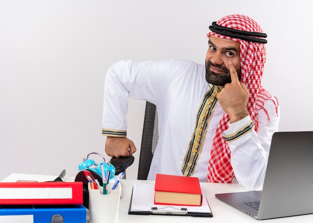 Arabische zakenman in traditionele slijtage zittend aan de tafel met laptop computerpointing met vinger aan zijn oog wachten op betaling werken in kantoor