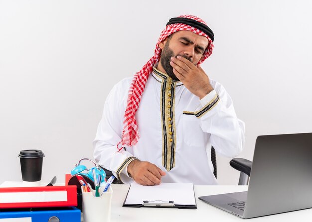 Arabische zakenman in traditionele slijtage zittend aan de tafel met laptop op zoek moe wil slapen geeuwen werken op kantoor