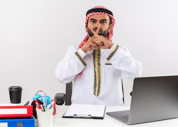 Gratis foto arabische zakenman in traditionele slijtage zittend aan de tafel met laptop overschrijden wijsvingers stilte gebaar werken in kantoor