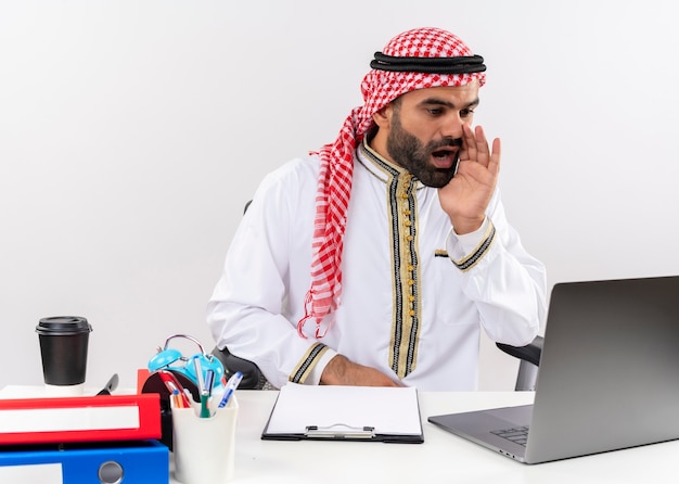 Arabische zakenman in traditionele slijtage zittend aan de tafel met laptop schreeuwen met hand in de buurt van mond werken in kantoor