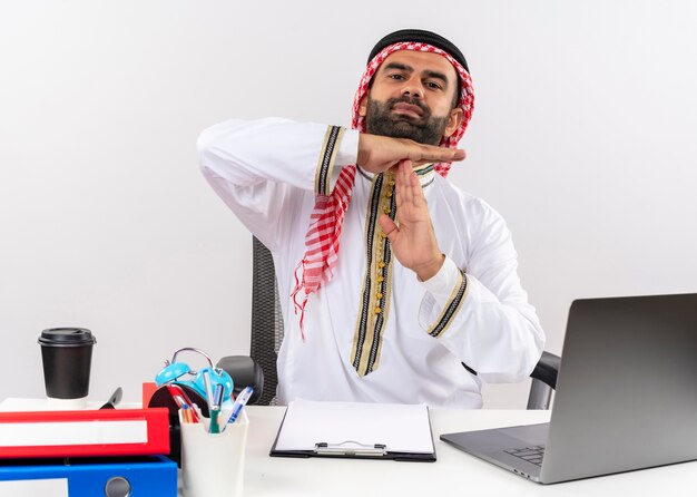 Arabische zakenman in traditionele slijtage zittend aan de tafel met laptopcomputer time-out gebaar maken met handen werken in kantoor gehinderd