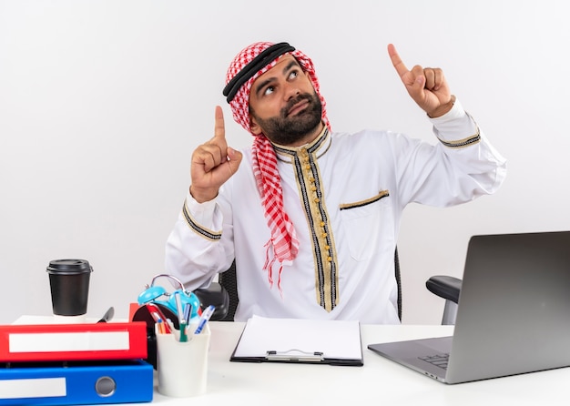 Arabische zakenman in traditionele slijtage zittend aan de tafel met laptopcomputer wijzend met wijsvingers op zoek zelfverzekerd werken op kantoor