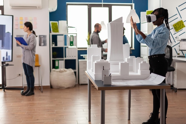 Architect staat in modern architectenbureau met behulp van virtual reality-bril om 3D-plan van witte schuimmaquette te bekijken. Ingenieur werkt met vr headest naast tafel met model van stedelijk project.