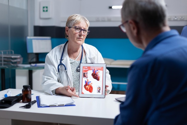 Arts die cardiologiediagnose met afbeelding van hartorgel op tablet aan patiënt uitlegt. Huisarts die illustratie op cardiovasculair systeem en anatomische structuur toont aan de mens.