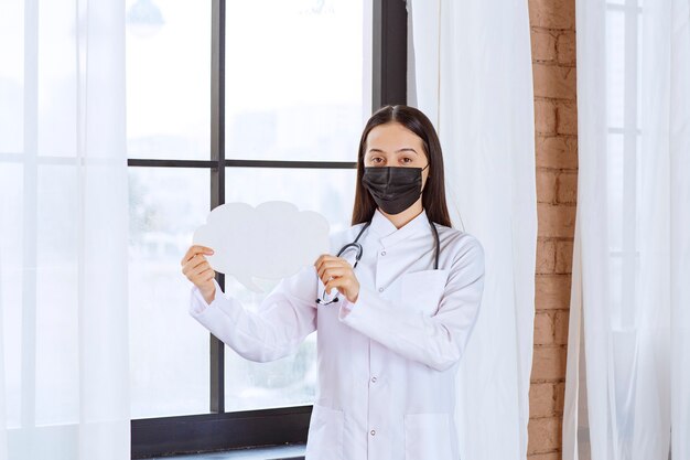 Arts die met een stethoscoop en een zwart masker een wit denkbord van de wolkenvorm houdt.