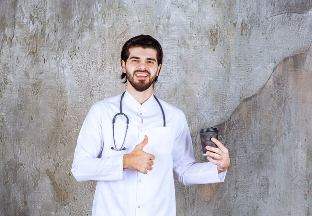 Arts met een stethoscoop die een zwarte wegwerpbeker vasthoudt en geniet van het moment of het product.