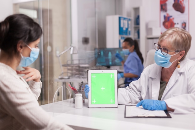 Arts met gezichtsmasker die tablet met groen scherm bekijkt tijdens overleg met zieke patiënt. Verpleegster die blauw uniform draagt.