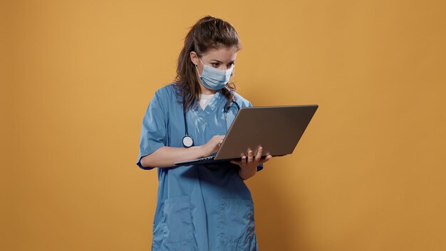 Arts met stethoscoop die laptop vasthoudt en op toetsenbord typt en naar het scherm kijkt dat in de studio staat. Dokter in ziekenhuisuniform met chirurgisch masker met draagbare computer.