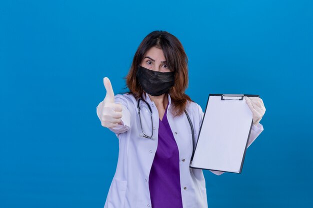 Arts van middelbare leeftijd, gekleed in een witte jas in een zwart beschermend gezichtsmasker en met een stethoscoop met klembord met lege plekken die er positief uitzien en duimen opdagen over blauwe muur