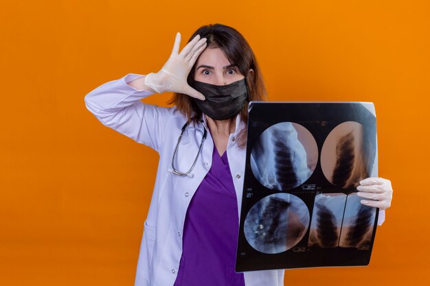 Arts van middelbare leeftijd met witte jas in zwart beschermend gezichtsmasker en met een stethoscoop met röntgenfoto van de longen op zoek verrast met de hand in de buurt van het hoofd over de oranje muur