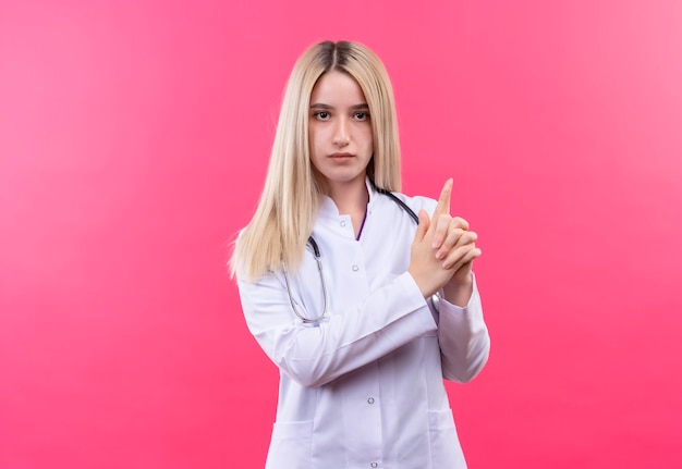 artsen jong blond meisje die stethoscoop in medische toga dragen die pistoolgebaar op geïsoleerde roze muur tonen