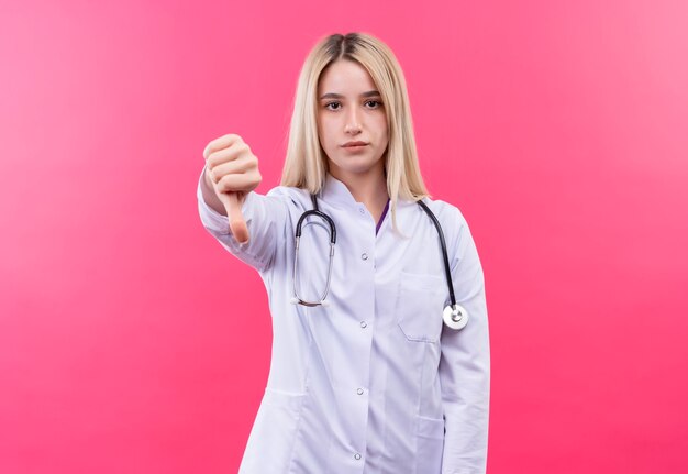 artsen jong blond meisje die stethoscoop in medische toga dragen haar duim neer op geïsoleerde roze muur