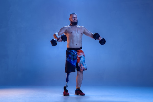 Atleet met een handicap of geamputeerde geïsoleerd op blauwe muur. professionele mannelijke sportman met beenprothese training met gewichten in neon. gehandicapte sport en overwinnen, wellness-concept.