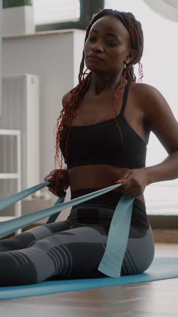 Atleet met zwarte huid in sportkleding die lichaamsspier uitoefent met behulp van fitnesselastiek en geniet van een gezonde levensstijl zittend op een yogakaart in de woonkamer