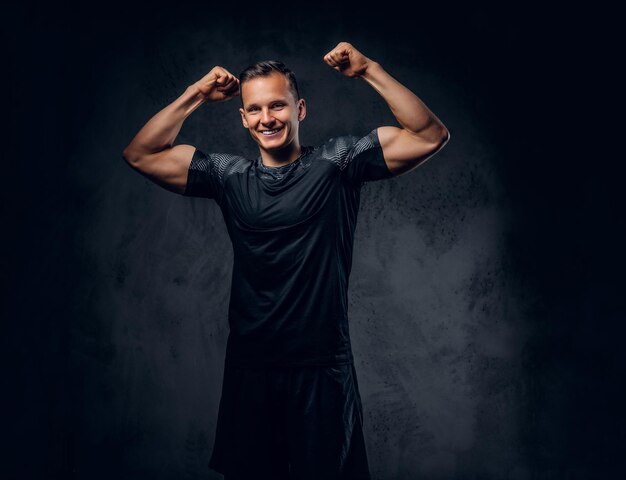 Atletische man gekleed in een donkere sportkleding toont zijn biceps op een grijze achtergrond.
