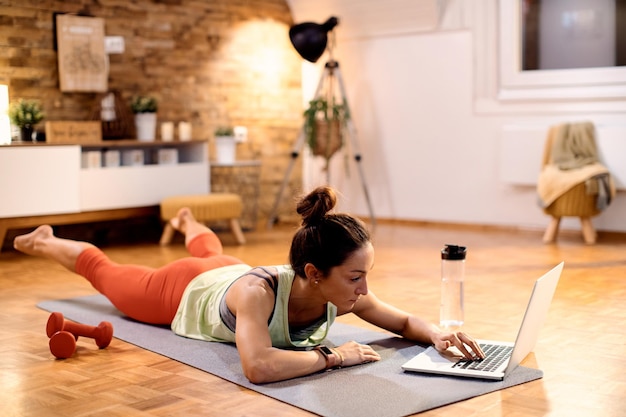 Atletische vrouw die laptop gebruikt terwijl ze thuis oefeningen op de vloer doet