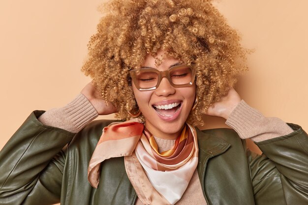 Authentiek emotiesconcept. Blije vrouw met krullend haar glimlacht breed houdt ogen gesloten glimlacht vreugdevol draagt leren jas en hoofddoek geïsoleerd over beige achtergrond lacht vrolijk om camera