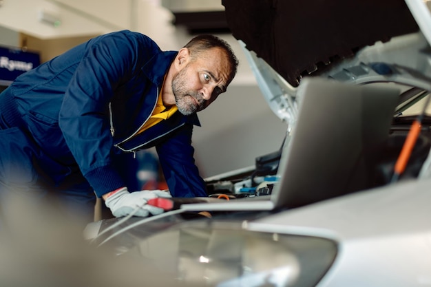 Gratis foto automonteur die laptop gebruikt terwijl hij de motor in de autoreparatiewerkplaats onderzoekt