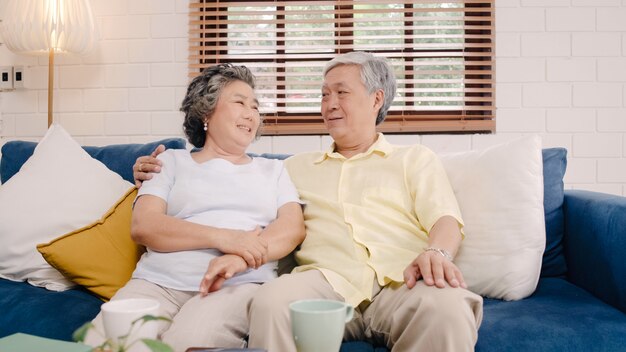 Aziatisch bejaard paar die het gelukkige glimlachen voelen en aan camera kijken terwijl op de bank in woonkamer thuis ontspant.