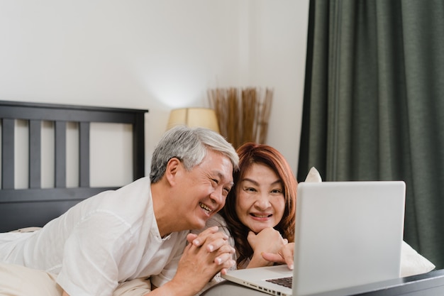 Aziatisch hoger paar dat laptop thuis met behulp van. Aziatische Hogere Chinese grootouders, echtgenoot en vrouw gelukkig na kielzog omhoog, lettend op film liggend op bed in slaapkamer thuis in het ochtendconcept.