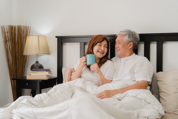Aziatisch hoger paar dat op bed thuis spreekt. De Aziatische Hogere Chinese grootouders, echtgenoot en vrouw drinken gelukkig koffie na kielzog omhoog terwijl thuis het liggen op bed in slaapkamer in het ochtendconcept.