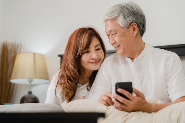 Aziatisch hoger paar die mobiele telefoon thuis met behulp van. Aziatische Hogere Chinese grootouders, echtgenoot en vrouw gelukkig na kielzog omhoog, lettend op film liggend op bed in slaapkamer thuis in het ochtendconcept.