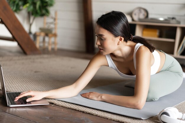 Aziatisch meisje doet yoga die zich uitstrekt thuis training in haar woonkamer kijken naar laptop scherm kijken...