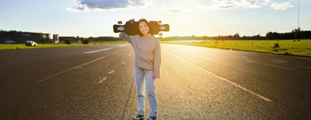 Gratis foto aziatisch meisje met skateboard staat op de weg tijdens zonsondergang skater poseert met haar lange board cruiser