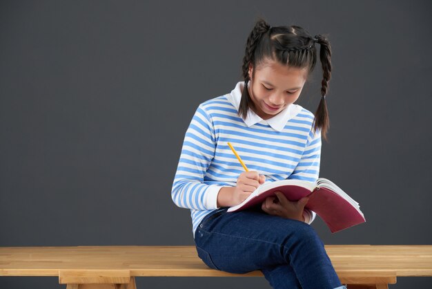 Aziatisch schoolmeisje met vlechten die op lijst zitten en in notitieboekje schrijven