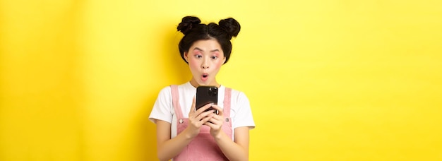 Aziatisch tienermeisje kijkt opgewonden naar het scherm van de smartphone en leest nieuws op de telefoon die op gele pagina staat