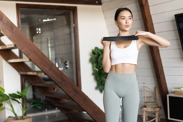 Aziatische fitness meisje doet training met elastische band uitrekken, oefenen voor sterke en fit armen, biceps en spieren, staande in activewear thuis in de woonkamer