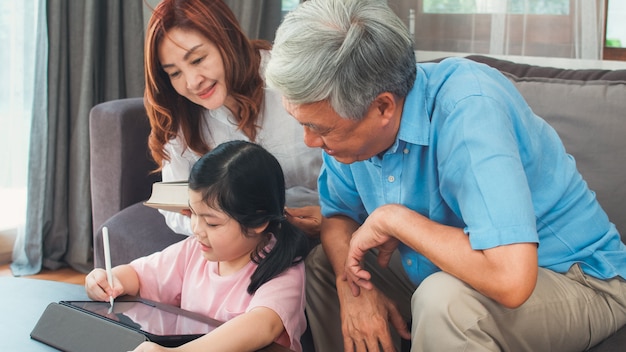 Aziatische grootouders en kleindochter videogesprek thuis. Hogere Chinees, opa en oma gelukkig met meisje die mobiel telefoonvideogesprek met behulp van die met papa en mamma spreken die in woonkamer thuis liggen.