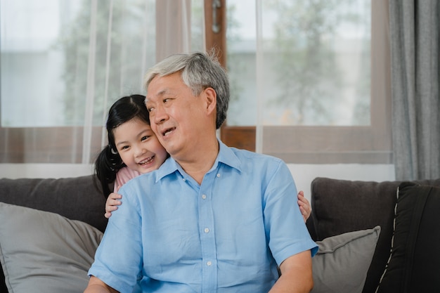 Aziatische grootvader die met kleindochter thuis spreekt. Hogere Chinees, gelukkig opa ontspant met jong kleindochtermeisje die familietijd gebruiken ontspant met jong meisjesjong geitje liggend op bank in woonkamer.