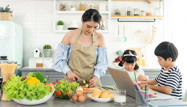 Aziatische jonge alleenstaande moeder die voedsel maakt terwijl ze voor een kind zorgt in het concept van de thuisschool in de keuken