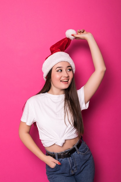 Aziatische jonge vrouw die de hoed van de Kerstman draagt die op roze muur wordt geïsoleerd