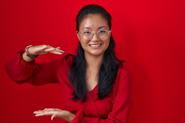 Aziatische jonge vrouw die zich over rode achtergrond bevindt die met handen gebaart die groot en groot teken maatsymbool glimlachen terwijl ze naar het meetconcept van de camera kijken