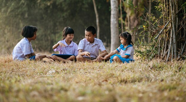 Aziatische jongen en drie kleine meisjes in studentenuniform zittend op gras en genieten van samen een handspel te spelen, ze praten en lachen met grappige, kopieerruimte, landelijk levensstijlconcept