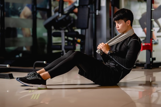 Aziatische knappe man met sportkleding en smartwatch zittend op de vloer, sit-up om spieren op te warmen voordat hij gaat trainen in de fitnessgym