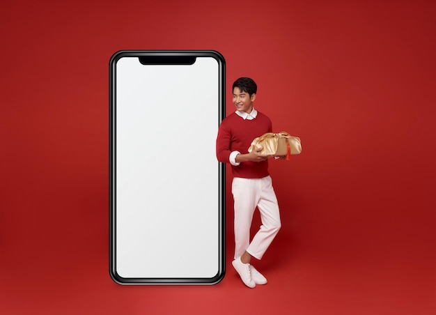 Aziatische man draagt een rode trui, houdt een cadeautje vast en kijkt naar het grote lege smartphone-scherm.