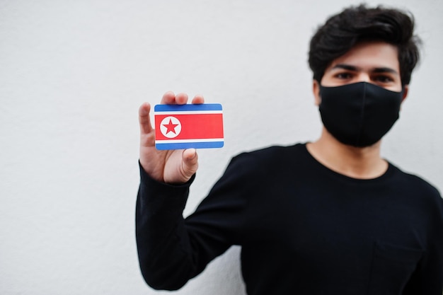 Aziatische man draagt helemaal zwart met gezichtsmasker houdt de vlag van noord-korea in de hand geïsoleerd op een witte achtergrond coronavirus landconcept