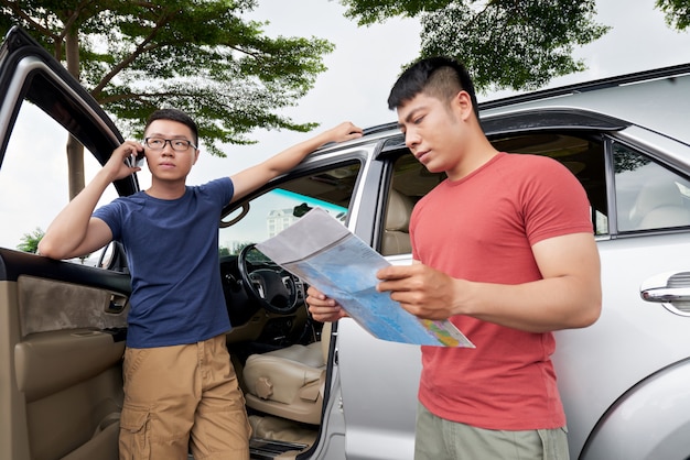 Aziatische mens die zich door zijn auto bevindt en op telefoon en vriend spreekt die kaart controleert