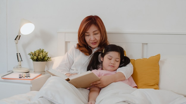 Aziatische oma las thuis sprookjes voor kleindochter. Hogere Chinees, gelukkige oma ontspant met jong meisje dat terwijl het luisteren aan verhalen thuis liggend op bed in slaapkamer thuis bij nachtconcept.